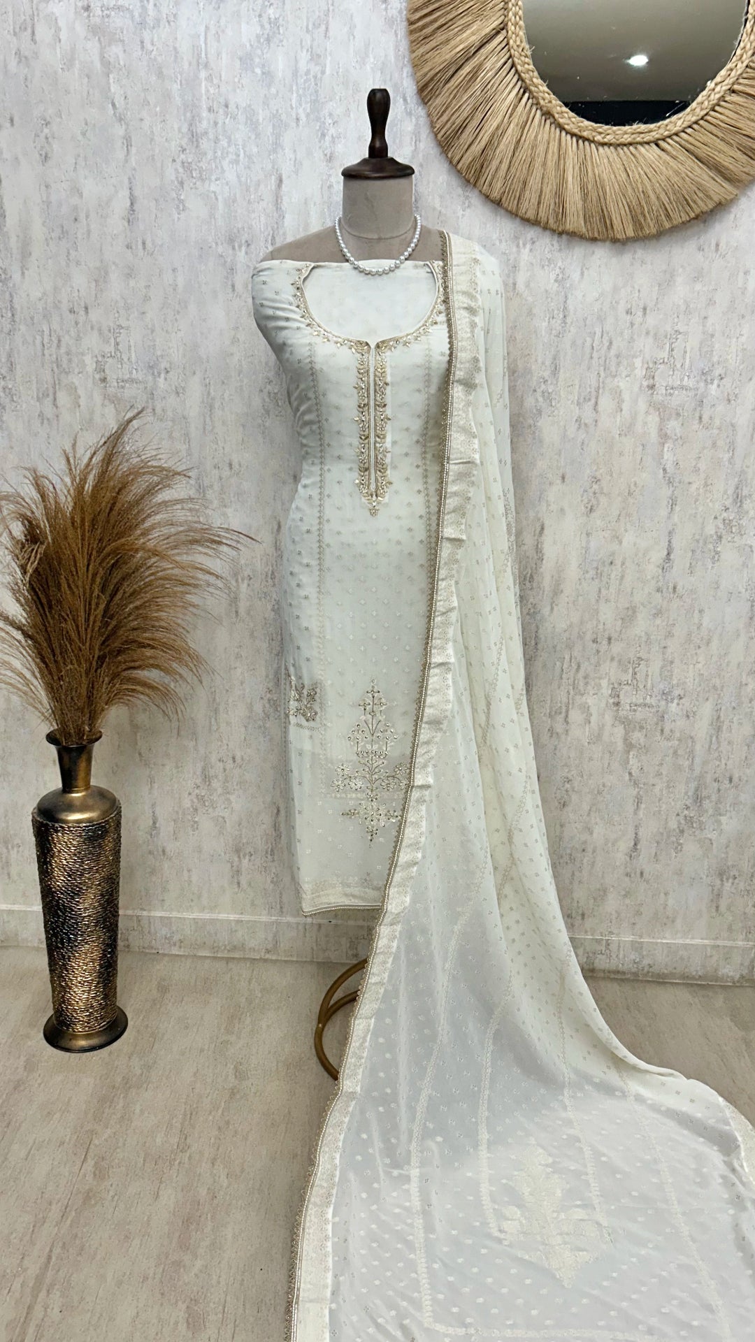 White embroidered dress on a mannequin with a decorative mirror and vase in the background.
